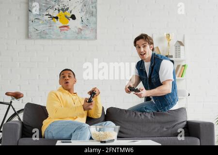 KYIV, UKRAINE - MARCH 22, 2021: interracial friends concentrating on playing video game with joysticks in modern living room Stock Photo
