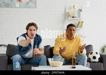 multiracial friends with emotional gestures watching game, drinking beer, eating popcorn and sitting on couch at home Stock Photo