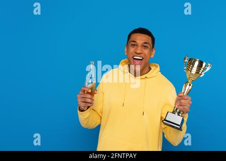 smiling african american man holding trophy cup and beer bottle in hands isolated on blue Stock Photo