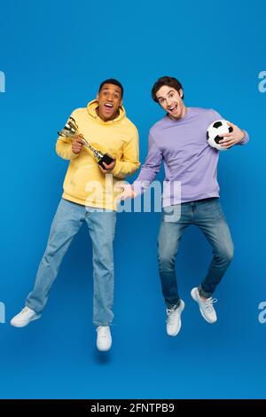 happy african american man with friend levitating and holding football and cup on blue background Stock Photo