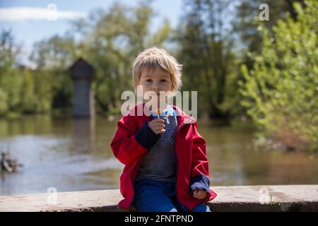 Child, sitting in front of old wooden cottage on brick pillar, eating lolly pop. Small cabin in Radbuza River, near Chotesov in Czech Republic, Europe Stock Photo