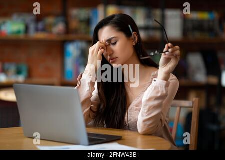Overworked latin freelancer woman tired after using laptop, working remotely on computer in cafeteria Stock Photo