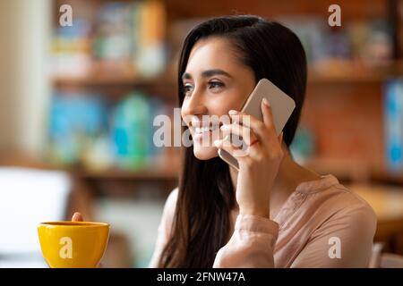 Happy latin lady talking on smartphone while drinking coffee and spending time alone at cafeteria Stock Photo