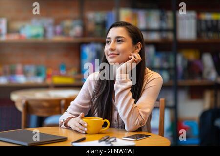 Dreamy arab lady using laptop at cafe, drinking coffee and dreaming, studying or working distantly Stock Photo