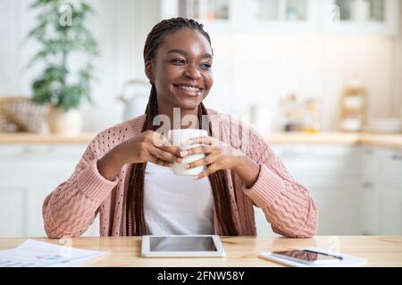 Relaxed Black Woman Sitting At Table In Kitchen And Drinking Coffee Stock Photo
