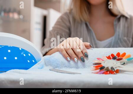 Woman with artificial acrylic nails picks up new polish color during manicure procedure. Manicure process in beauty salon. Hygiene beauty of hands in Stock Photo