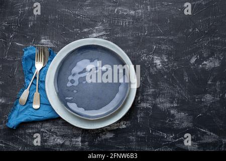 Empty blue plate served with fork spoon table napkin. Mockup template vintage blue plate for luxury dinner in restaurant. Dark black concrete table Stock Photo