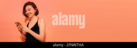 young woman looking at smartphone in hands and smiling on orange background, banner Stock Photo