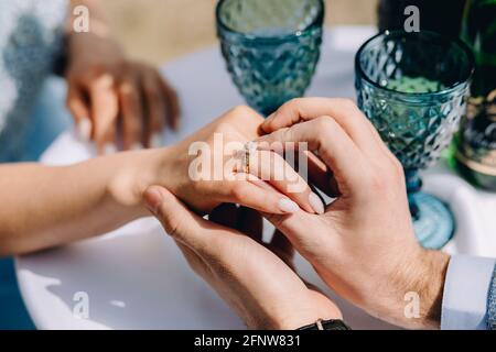 Outdoor close up of man proposing to his girlfriend. Guy puts engagement ring and leaning elbows on table with glasses Stock Photo