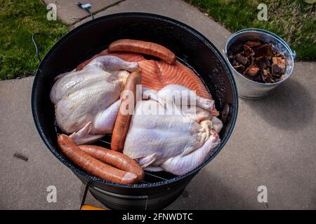 Barrel home food smoker sitting on sidewalk with two whole chickens and salmon and sausages on top rack - soaking wood sitting in bucket nearby Stock Photo