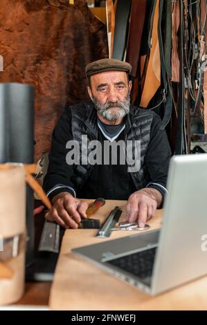 Senior man working with leather in his workplace and looking at the camera with computer laptop. handicrafts products Stock Photo