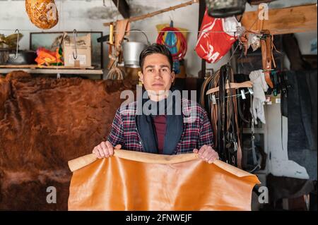 Young craftsman holding a piece of yellow genuine leather and looking at the camera. Guy working in his workshop. Stock Photo