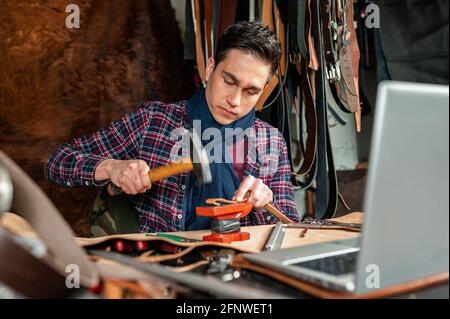 Young adult working with leather and holding a hammer while looking a tutorial video on computer laptop. Home hobbies and handicraft concept. Stock Photo