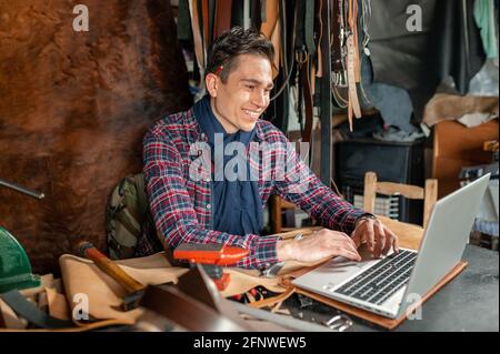 Smiling young artisan man sitting in his workplace while using computer laptop to learn how to work with leather. Home hobbies concept. Stock Photo