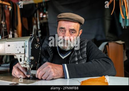 Old bearded man with serious face looking at the camera while using sewing machine working with leather. Stock Photo