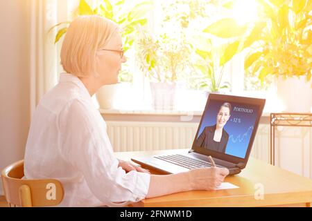 Senior woman at home in front of her laptop watching an online video of a female financial consultant. Stock Photo