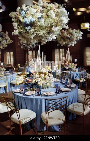 Round dining tables, covered in d blue cloth, served with porcelain and blue and purple glasses and richly decorated with flowers, flowers under the Stock Photo