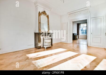 Vintage mirror placed on wooden decorative fireplace in spacious hallway with white walls with classic stucco elements and parquet floor Stock Photo