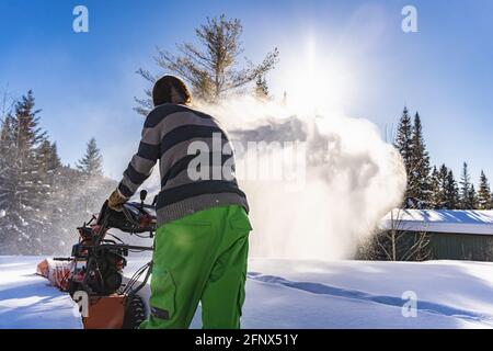 Close up shot of a yong man in yellow fluo pants digging a path in the snow with an orange mechanical snowplough in a sunny winter day in Canada. Stock Photo