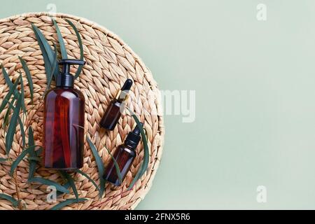 Dropper bottle of essential oil or serum, eucalyptus branches, and dark dispenser bottle of shower gel on rattan background. Beauty and SPA concept. Stock Photo