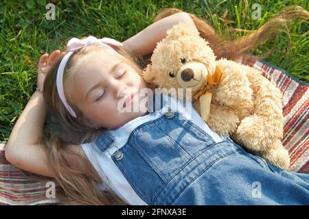 Pretty little child girl with closed eyes laying down with her teddy bear toy on blanket on green grass in summer taking a nap. Stock Photo