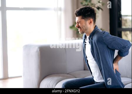 Unhealthy sad young adult man stand up from couch in living room touches back, suffer from back pain. Caucasian guy suffers from arthritis, radiculitis, backache pain Stock Photo