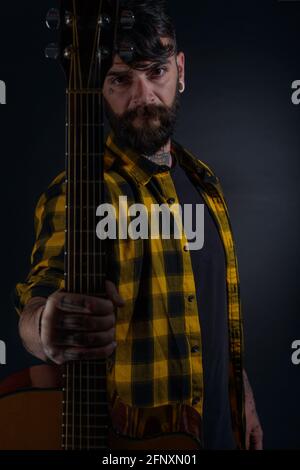 Tattooed man with a beard wearing a tartan yellow black shirt is standing and posing with his guitar in his hand Stock Photo