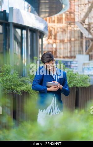 Well suited buisnessman is having a conversation on his phone with his partners while working on his tablet, in front of his company Stock Photo