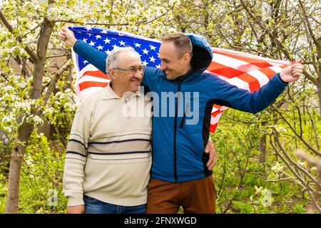 Handsome dad with his little cute sun are laying on green grassy lawn on American flag with American football ball in hand. Stock Photo