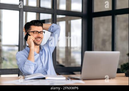 Smartphone conversation. Satisfied friendly caucasian man, office worker or entrepreneur, sitting at a table in the office, having a pleasant phone conversation while working, smiling Stock Photo