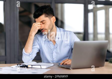 Headache and migraine. Exhausted caucasian young adult man, manager, freelancer or office employee, tired of works, in stress, took a break from work, need a rest, massages eyes, have a headache Stock Photo