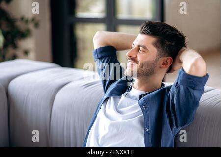 Closeup of happy bearded caucasian adult man in casual clothes relaxing on couch at home. Relaxed guy having break on sofa in living room, dreaming about vacation, looks away, smiling. Stock Photo