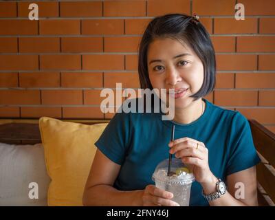 Portrait of Asian middle aged woman looking at camera, holding ice berverage.  Brick wall, sitting on sofa. Stock Photo