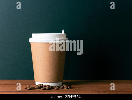Disposable paper cup with hot coffee and roasted coffee grains on wooden table in front of black wall. Space for text Stock Photo