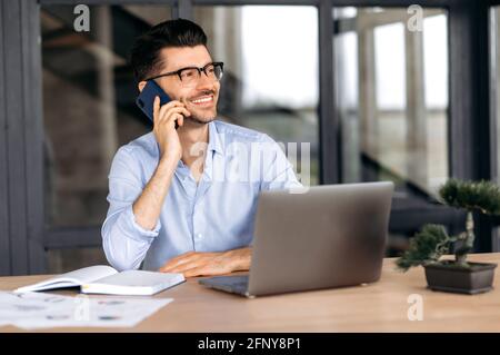 Mobile communication concept. Smiling caucasian freelancer talking on cellphone while working on laptop, positive man in formal shirt having pleasant phone conversation, smiling friendly Stock Photo