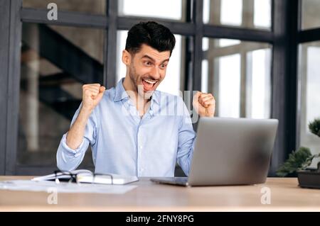 Happy excited caucasian male freelancer or office worker, sitting at his work desk at office, using laptop, shouts and rejoice in success, win or good profit, looking at laptop and gesturing hands Stock Photo