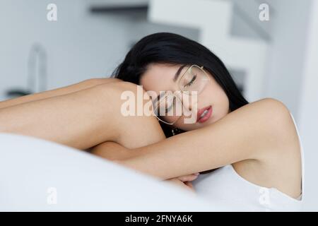 Tired young woman wearing trendy glasses sleeping peacefully clasping her knees as she relaxes on a white sofa in close up with parted lips and serene Stock Photo