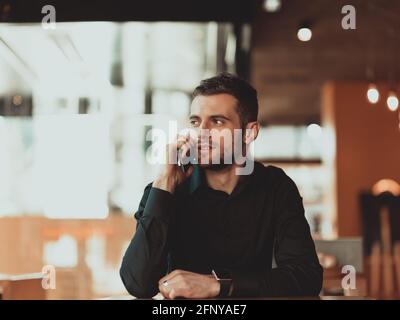 a young businessman in a black shirt sits in a coffee shop and uses smartphone during a coffee break. Stock Photo
