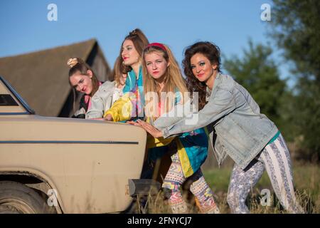 Young cheerful girls are pushing an old car. Women in the style of the 90s. Stock Photo