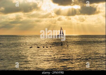 A Sailboat Is Silhouetted Against Sun Rays Beaming Down On The Ocean Horizon In Golden Shafts Of Light Stock Photo