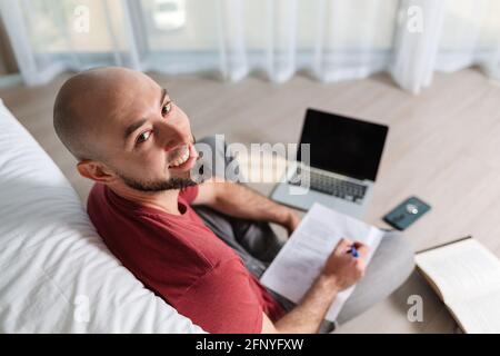 A bald young happy man is working at a laptop and writing something down on paper. Top view. The concept of online courses and online learning. Stock Photo