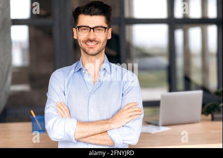 Formal business portrait. Confident successful caucasian businessman or manager with glasses stands near his work desk in the office, arms crossed, looks directly at the camera and smiles friendly Stock Photo