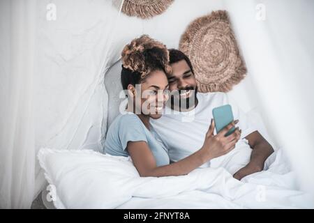 Darkskinned woman and man taking selfie in bed Stock Photo
