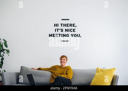Smiling businesswoman on sofa in lobby. Young female professional looking away and thinking in office lobby. Stock Photo