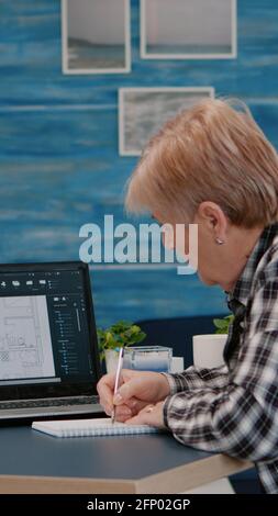 Senior woman architect analysing digital prototype with plans from laptop working from home, taking notes on notebook. Elderly designer using cad software to create a 3D concept of buildings Stock Photo