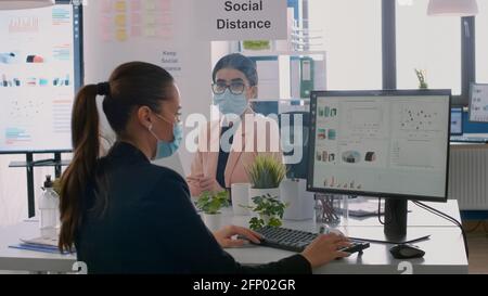 Collegues with protection medical face masks sitting at desk talking about business project in workplace office. Coworkers keeping social distancing to avoid virus disease during global epidemic Stock Photo