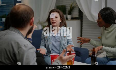 Mixed race friends socializing during entertainment party while playing guess who game attaching stickey notes. Multiracial people spending time together having fun during entertainment wekeend Stock Photo