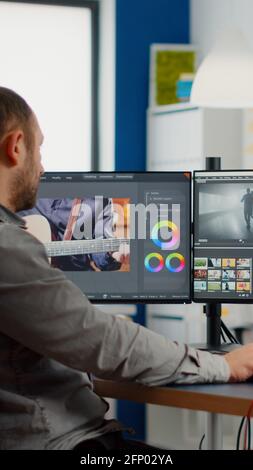 Videographer edits video project and cuts footage and sound using post production software and two monitors taking notes. Man processing film montage in digital multimedia star up company Stock Photo