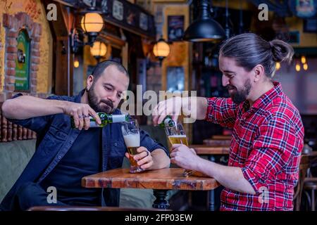 Two male friends drinking in the pub. Pouring beer from the bottle in tall glasses. Back to normal life. Stock Photo