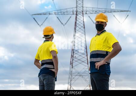 Electrical engineers working. Electrician looking the camera and another skilled worker looking high voltage tower against blue sky. Stock Photo
